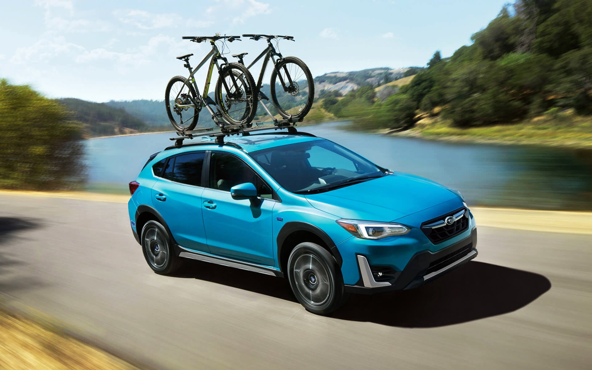 A blue Crosstrek Hybrid with two bicycles on its roof rack driving beside a river | Jim Keras Subaru in Memphis TN